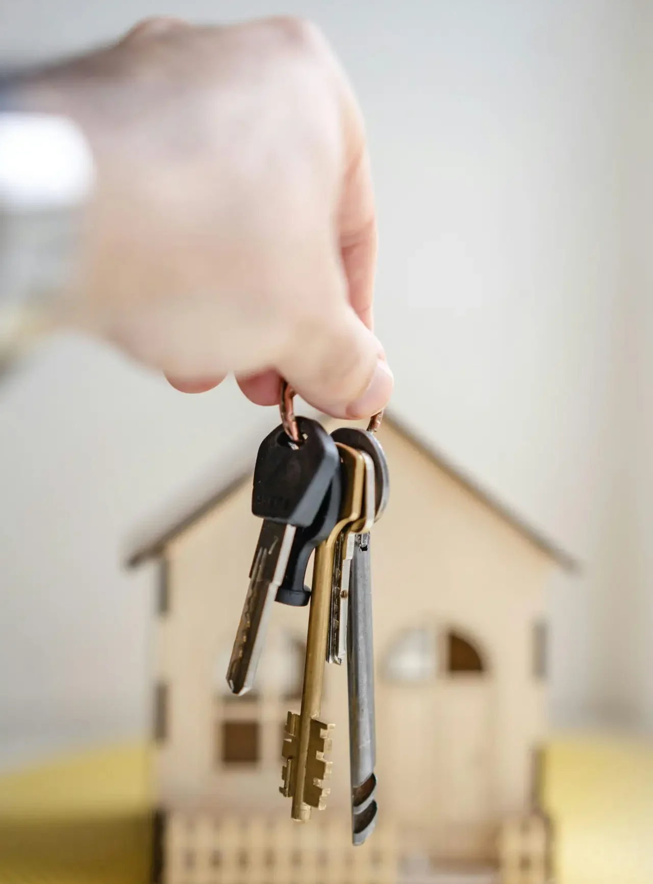 Close-up of a hand holding keys with a miniature wooden house in the background, symbolizing real estate investment.
